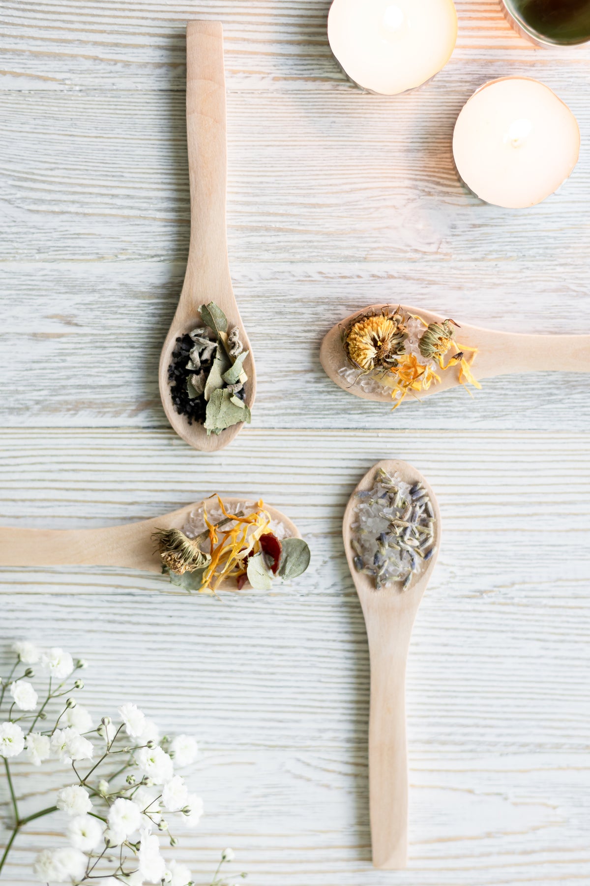 Cuatro cucharas de madera con sales y hierbas secas como caléndula, lavanda y hojas sobre superficie de madera blanca, con velas de té y flores blancas al borde