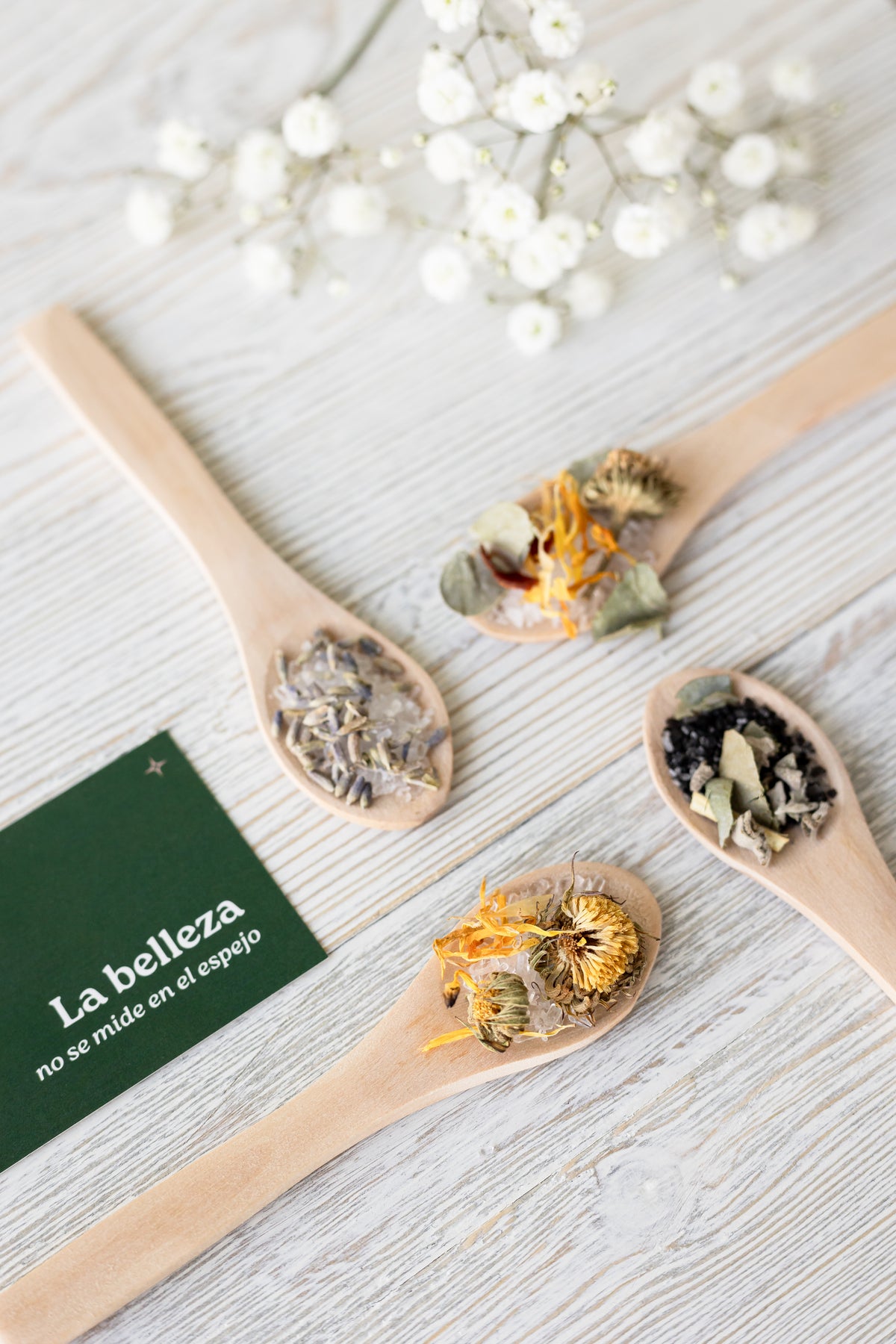 Cuatro cucharas de madera con sales de lavanda, caléndula y semillas sobre madera blanca, junto a una tarjeta verde que dice La belleza no se mide en el espejo y flores blancas