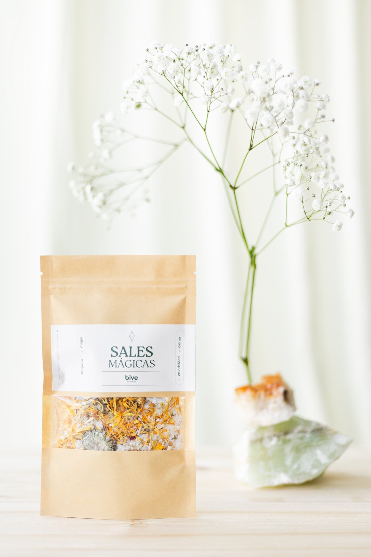 Sobre kraft de Sales Mágicas de Limpieza bive con ventana que muestra pétalos naranjas, junto a un jarrón con flores blancas paniculata y cristales