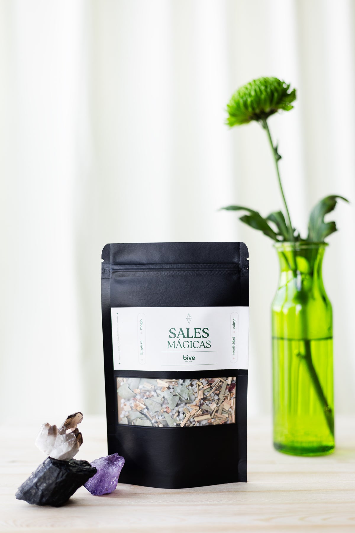 Sobre negro de Sales Mágicas de Limpieza bive con ventana que muestra la mezcla de sales, junto a un jarrón de vidrio verde con crisantemo verde y cristales