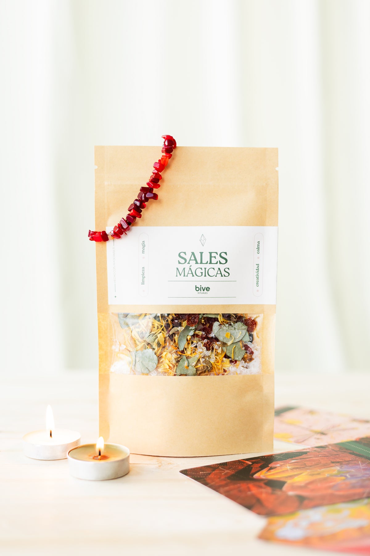 Sobre kraft de Sales Mágicas de Limpieza bive con pulsera de piedras rojas sobre el cierre, velas de té encendidas al frente y una tarjeta de arte colorida junto al sobre