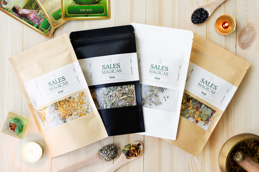 Composición plana de cuatro sobres de Sales Mágicas de Limpieza bive en kraft, negro y blanco sobre madera, rodeados de velas, cartas del tarot, cucharas con sales y hierbas y un mortero