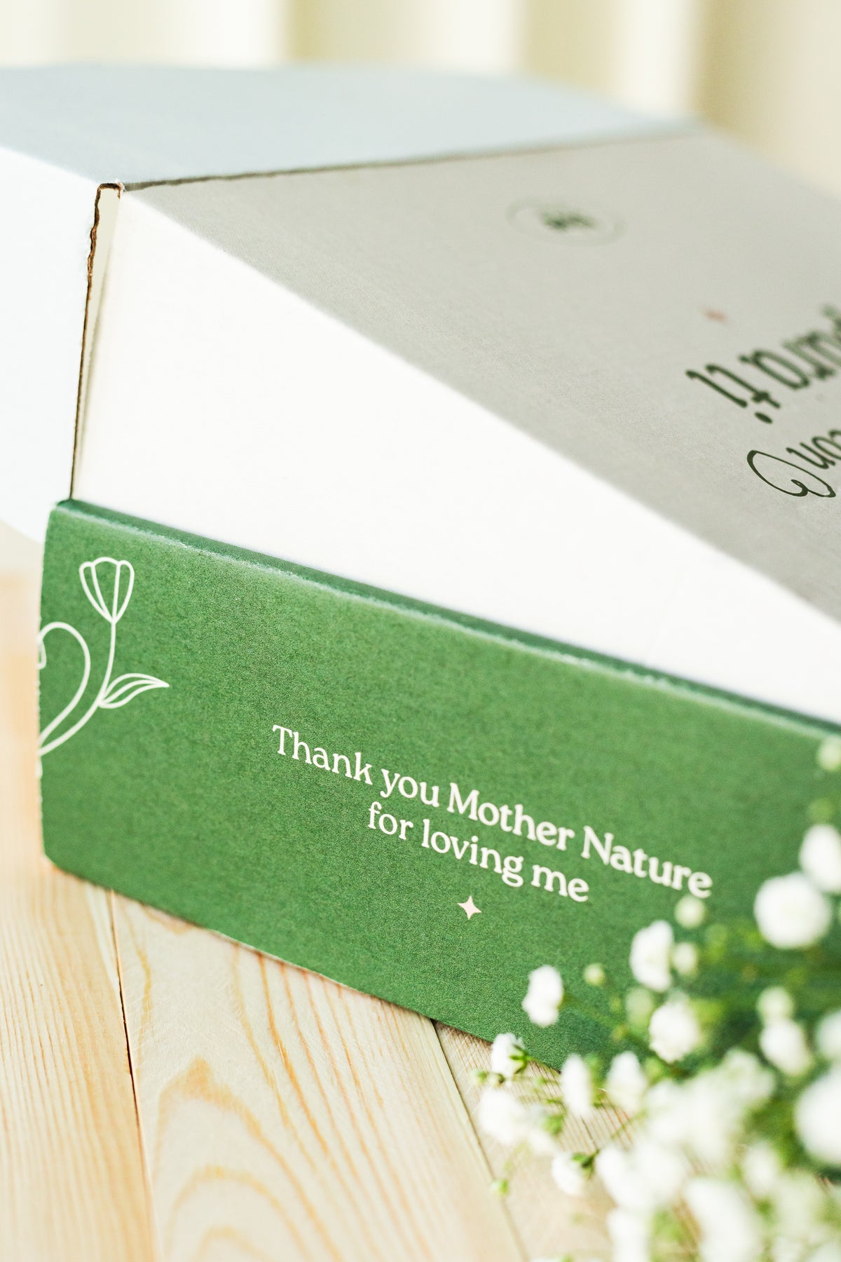 Caja verde de empaque con el texto Thank you Mother Nature for loving me sobre madera, con flores blancas paniculata