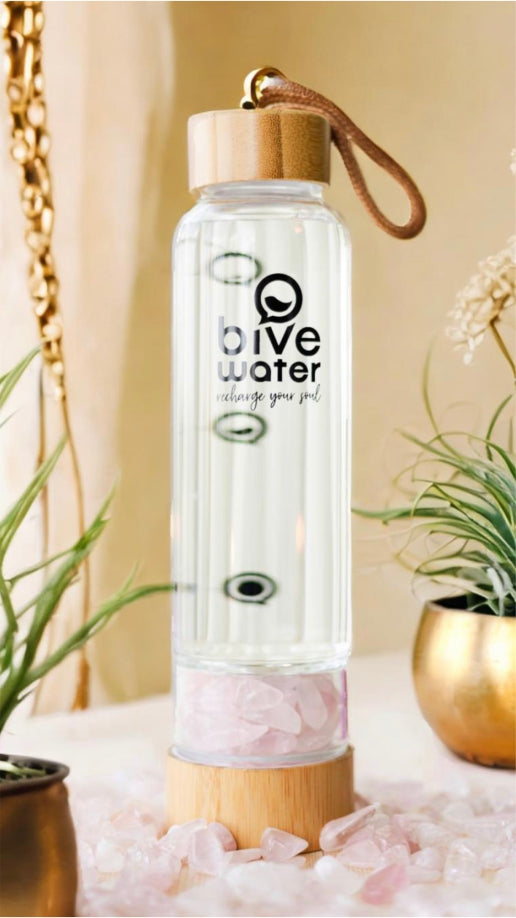 Botella alta de cristal Bive Water con tapa de bambú, con cuarzo rosa en su interior, sobre base de madera rodeada de piedras rosadas y plantas